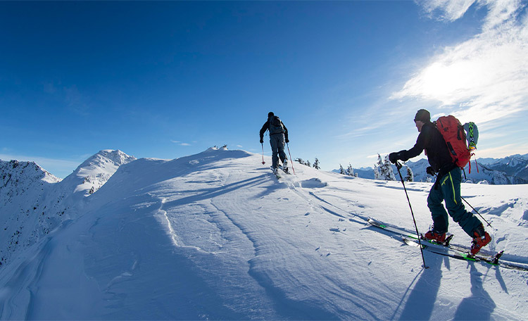 Door gericht skitoerentraining begin je fit aan het skitoerenseizoen. Twee toerskiërs beklimmen in de zon een besneeuwde berg.