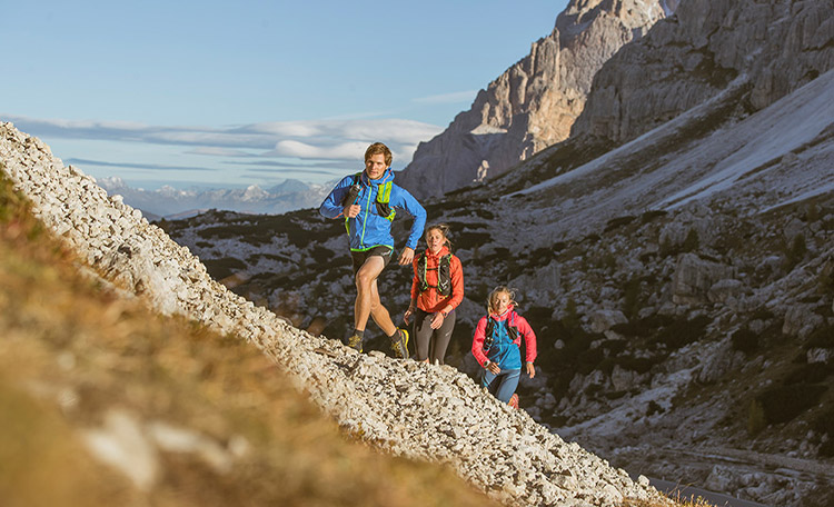 Hardlopen is bijzonder geschikt om stap voor stap de basisconditie te trainen. Drie trailrunners rennen in de zon een berg op.