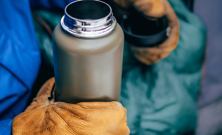 Warme slaapzak, dikke handschoenen en een thermoskan vol thee! Close-up van een thermosfles op de schoot van een warm geklede persoon.