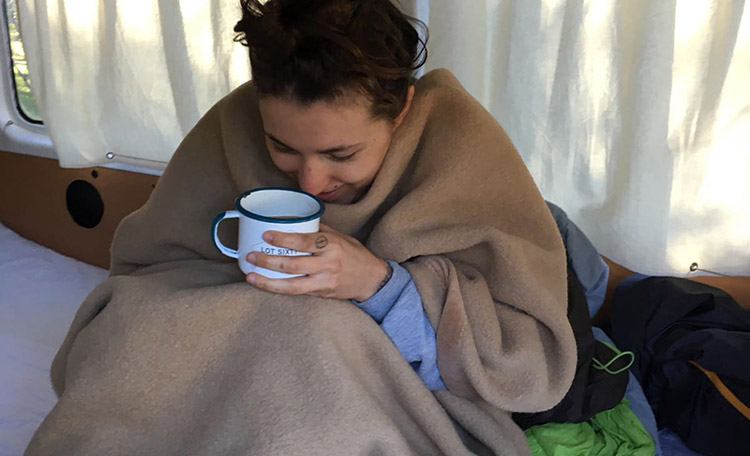 Heet thee, kruiken en dekens houden je warm in de campervan. Een vrouw nestelt zich in een campervan in een deken en heeft een kopje thee in haar hand.